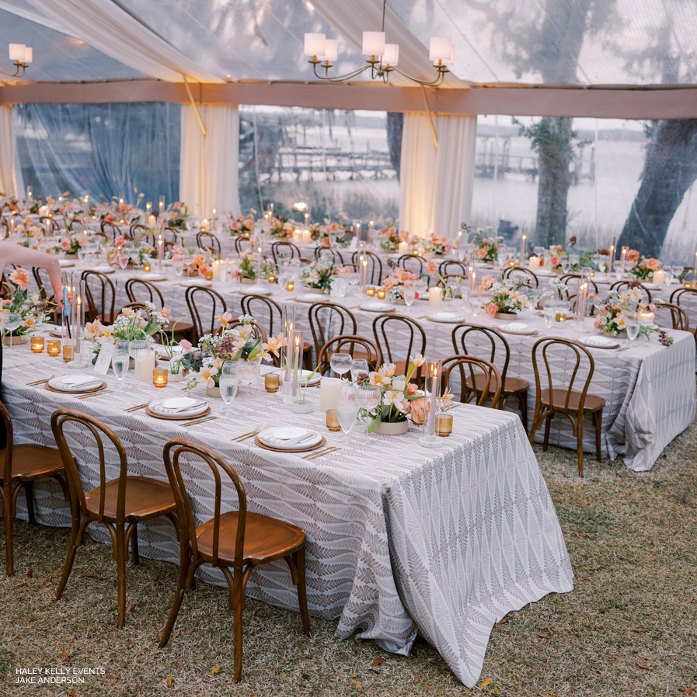 Elegantly set tables with Jolie Wheat centerpieces for an outdoor event under a tent with hanging lights.