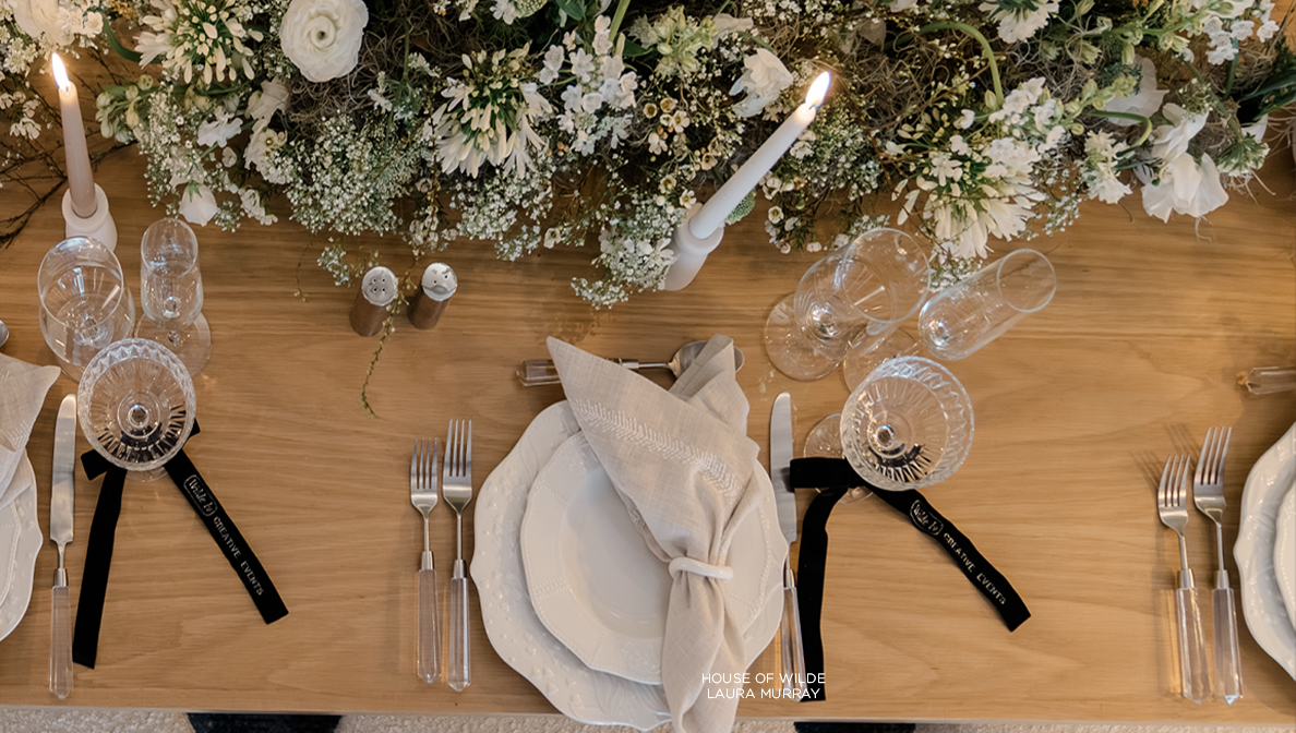 Elegant dining table setting featuring white flowers, greenery, crystal glasses, and black-handled cutlery with napkins folded on plates, perfect for a reverie social.
