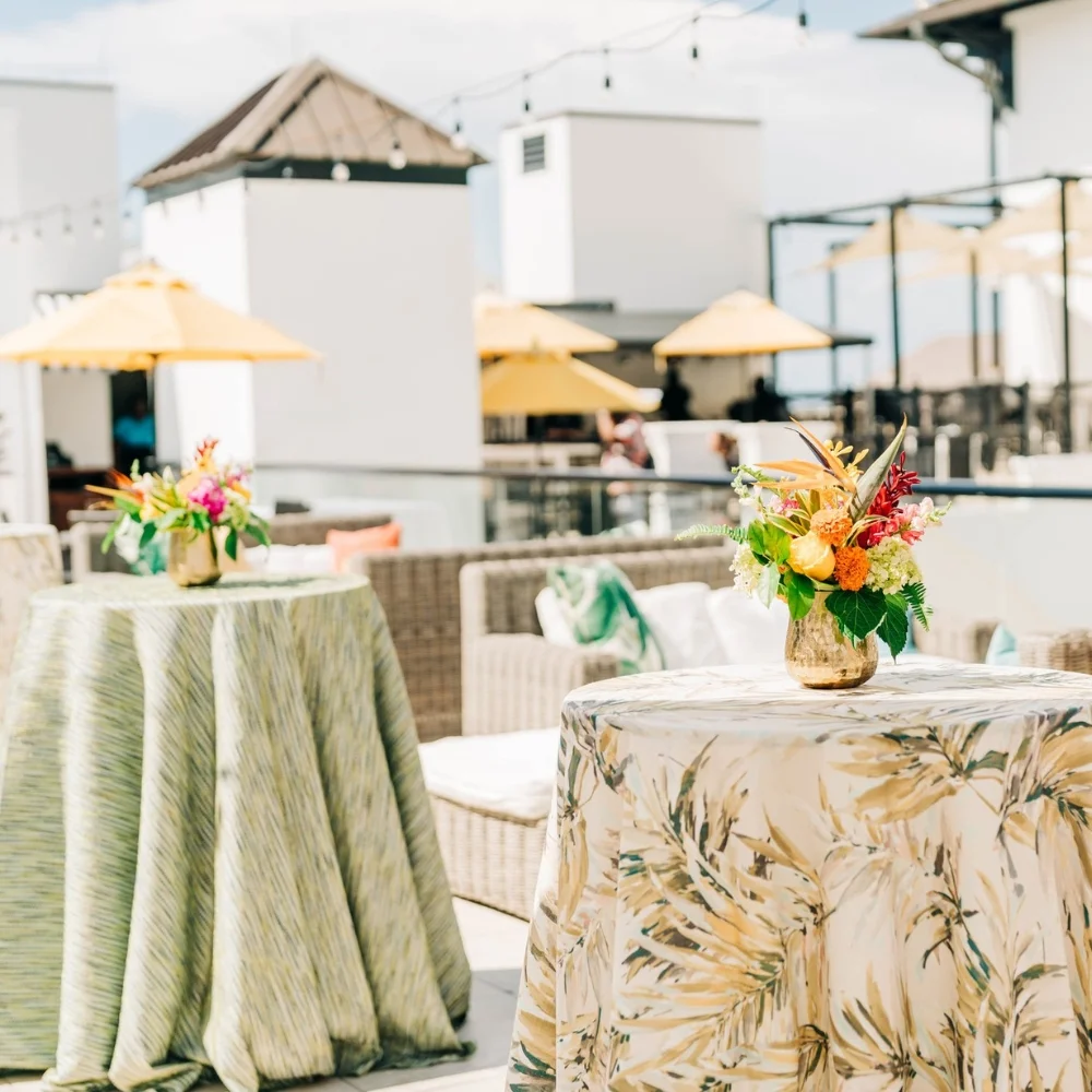 Outdoor rooftop event setup with tables covered in floral-patterned cloths, colorful flower arrangements, and yellow umbrellas in the background.