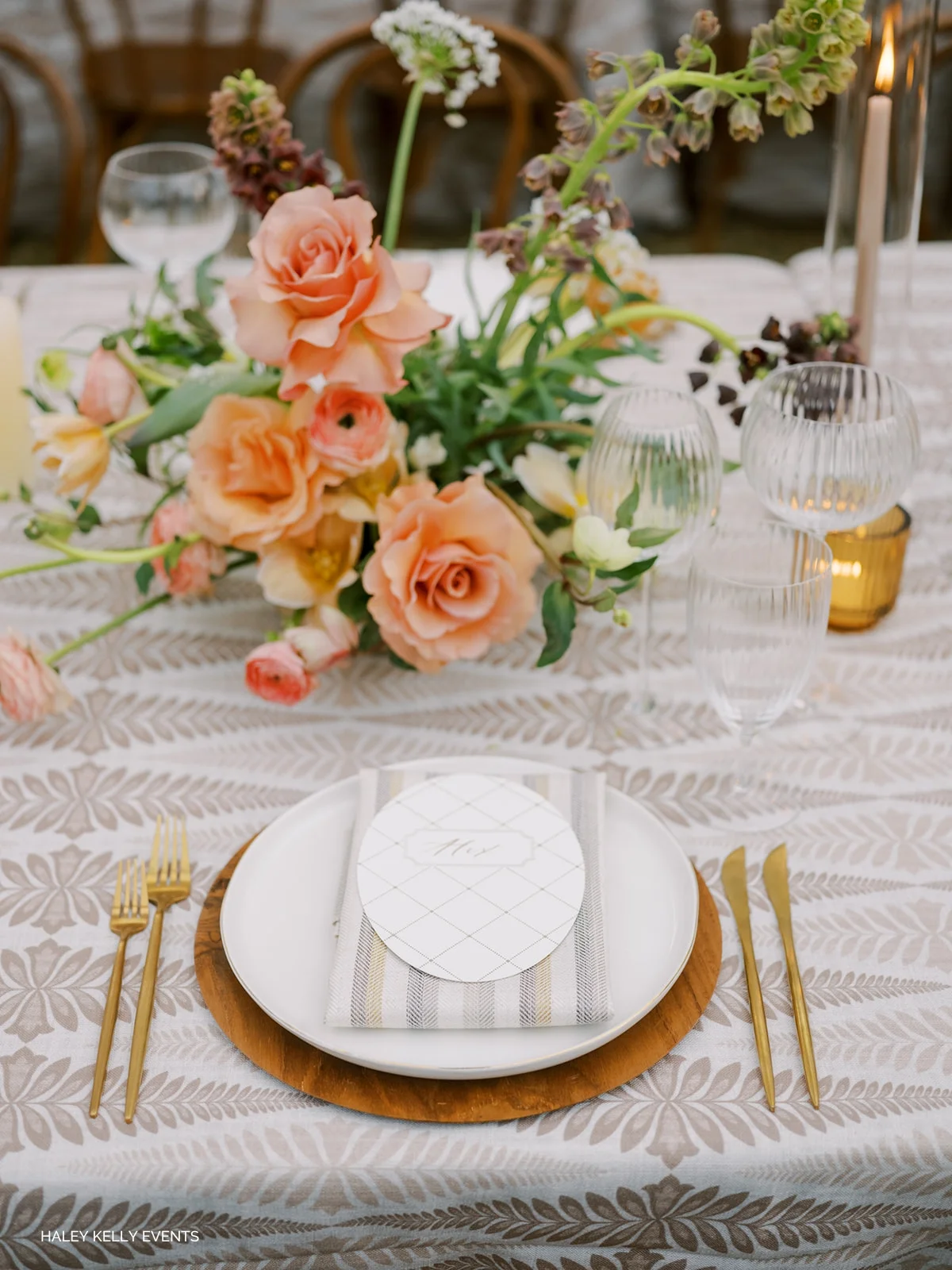 Elegant dining table setting featuring coral roses centerpiece, white plates, gold cutlery, and patterned tablecloth.