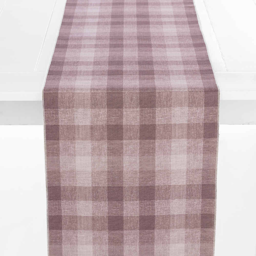 A Checkered Plaid Elderberry Table Runner with a mauve and white checkered pattern is laid on a plain white table.